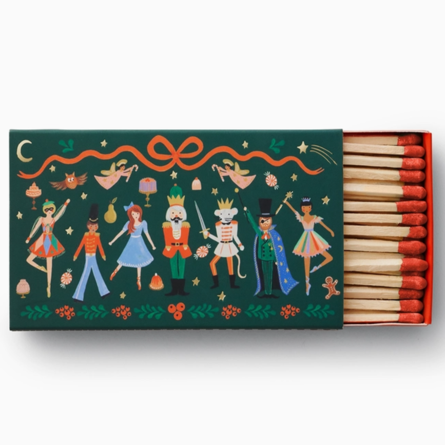  decorative matchbox with a festive theme. The box features illustrations of characters that appear to be inspired by "The Nutcracker" ballet, including a ballerina, a soldier, a mouse king, and other whimsical figures. The background is dark green with accents of orange, gold, and red, along with decorative ribbons, stars, and floral elements. The matchsticks inside have red tips