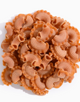 A close-up view of uniquely shaped pasta pieces in a vibrant reddish-brown color. The pasta features a curved design with ridges and frilly edges, resembling small mohawks.