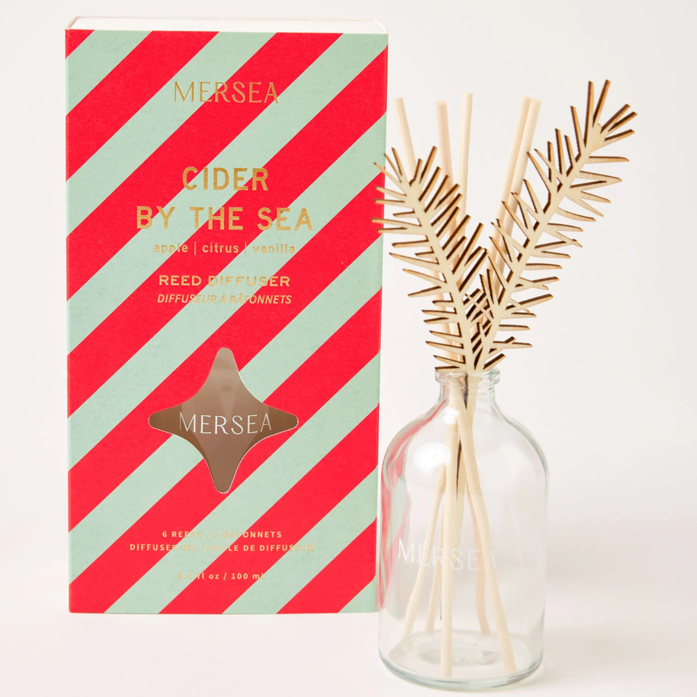 RED AND LIGHT BLUE STRIPED PACKAGING WRITTEN IN GOLD LETTERS: "MERSEA / CIDER BY THE SEA / APPLE, CITRUS, VANILLA / REED DIFFUSER" "6 WODDEN REEDS." NEXT TO IT THE GLASS DIFFUSER IS DISPLAYED WITH THE REEDS INSERTED INSIDE