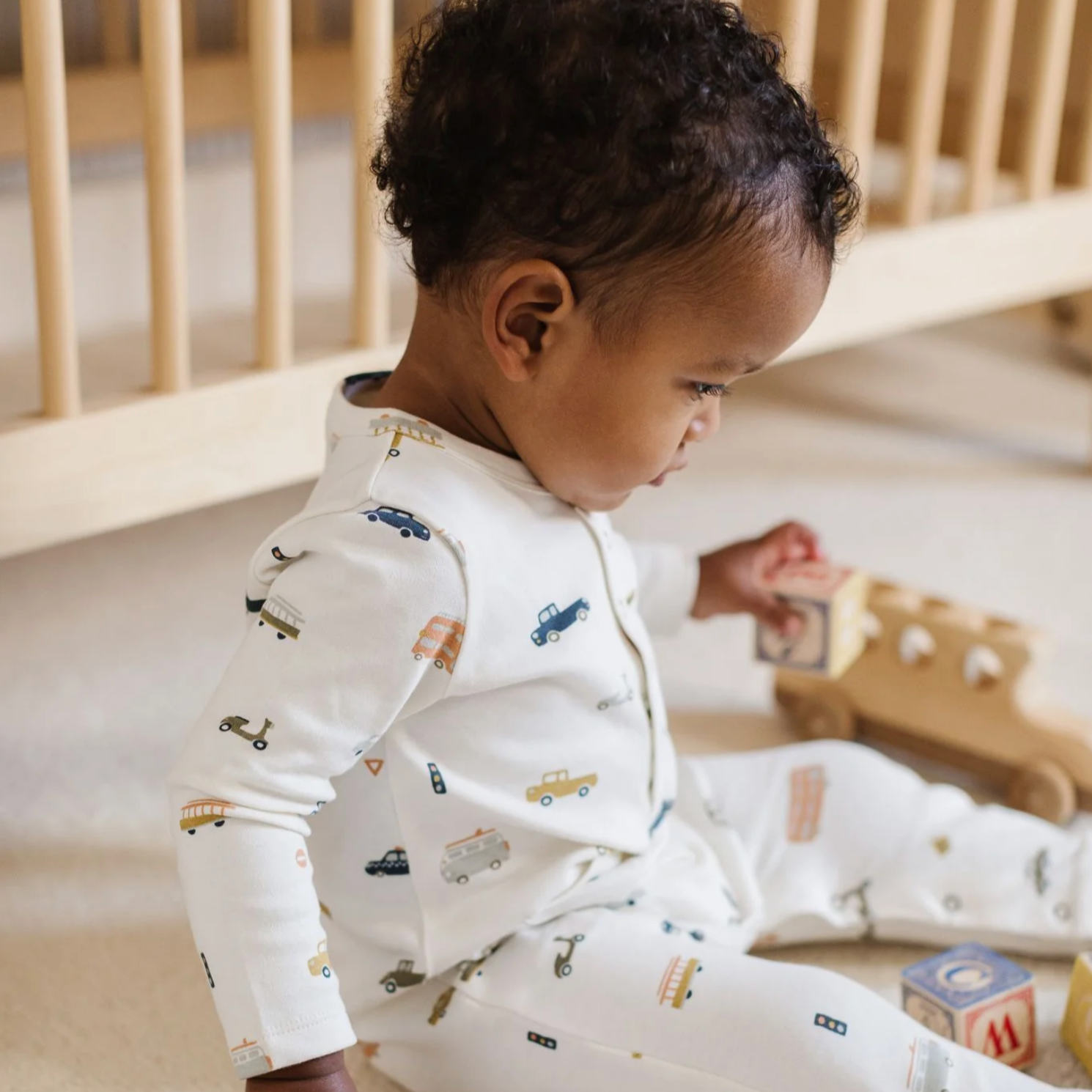 Baby in a white romper with multicolor car, bus and scooter print all over, metal snaps at collar and two pockets at hips.