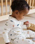 Baby in a white romper with multicolor car, bus and scooter print all over, metal snaps at collar and two pockets at hips.