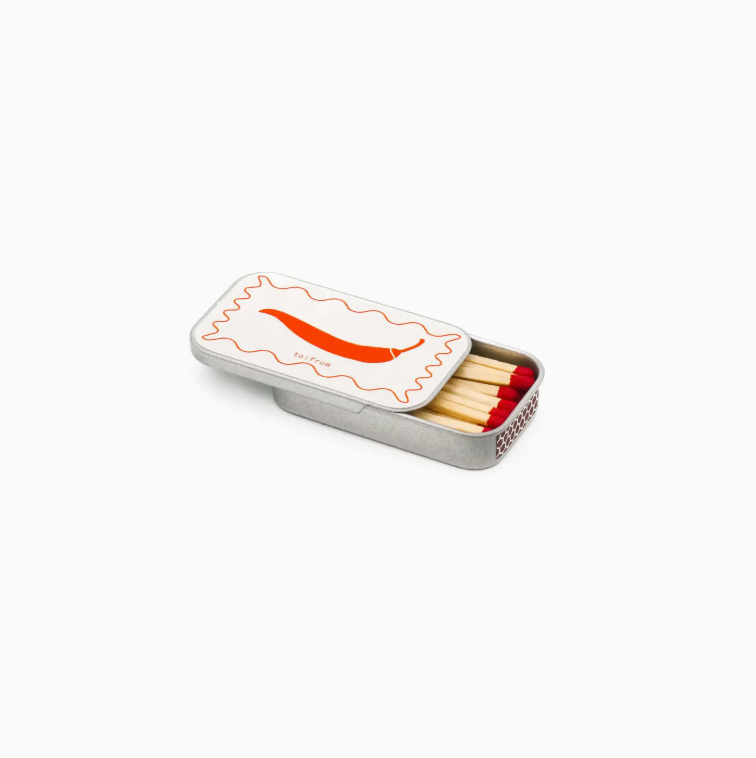 White background with half open mini tin matchbox in the center of the frame. Has red matches inside with orange chili pepper drawing and squiggle background on the front of the tin. 