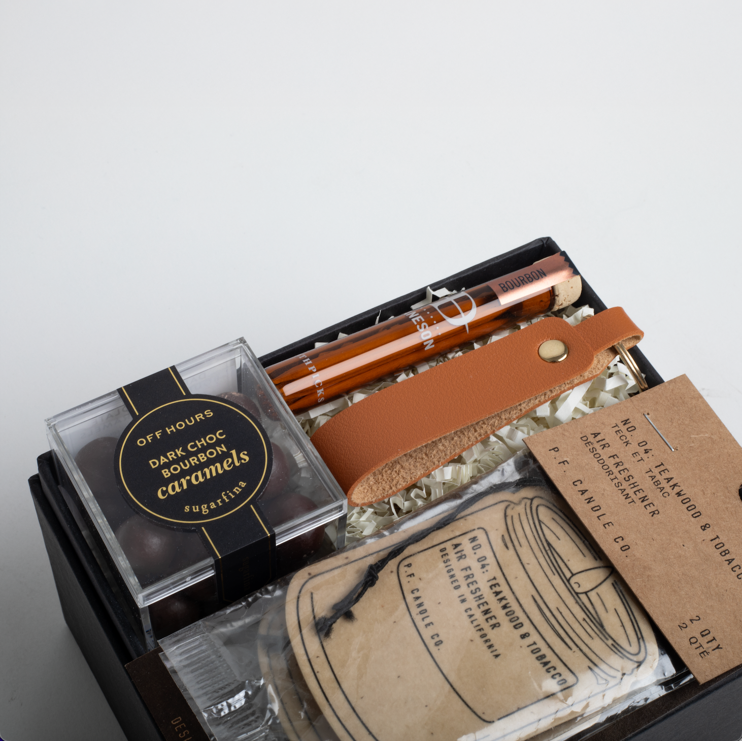 Gift set with dark chocolate bourbon caramels, a leather strap, and a jar of tobacco on a light gray background.