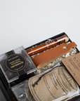 Gift set with dark chocolate bourbon caramels, a leather strap, and a jar of tobacco on a light gray background.