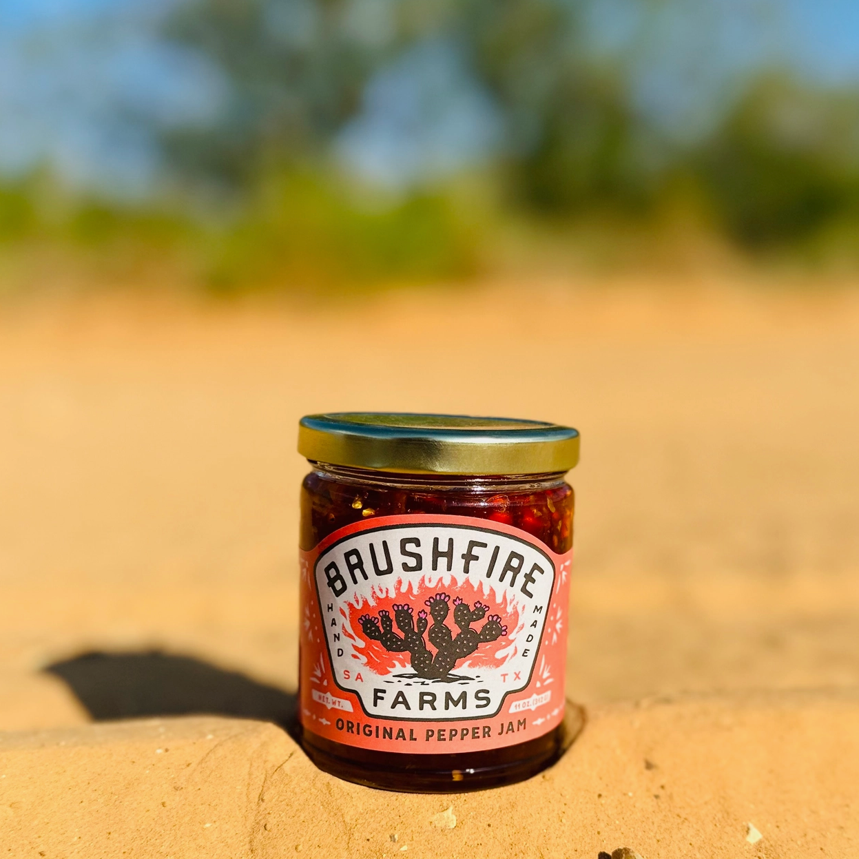 Jar of Bushfire Farms Original Pepper Jelly on a dirt surface with blurred greenery in the background