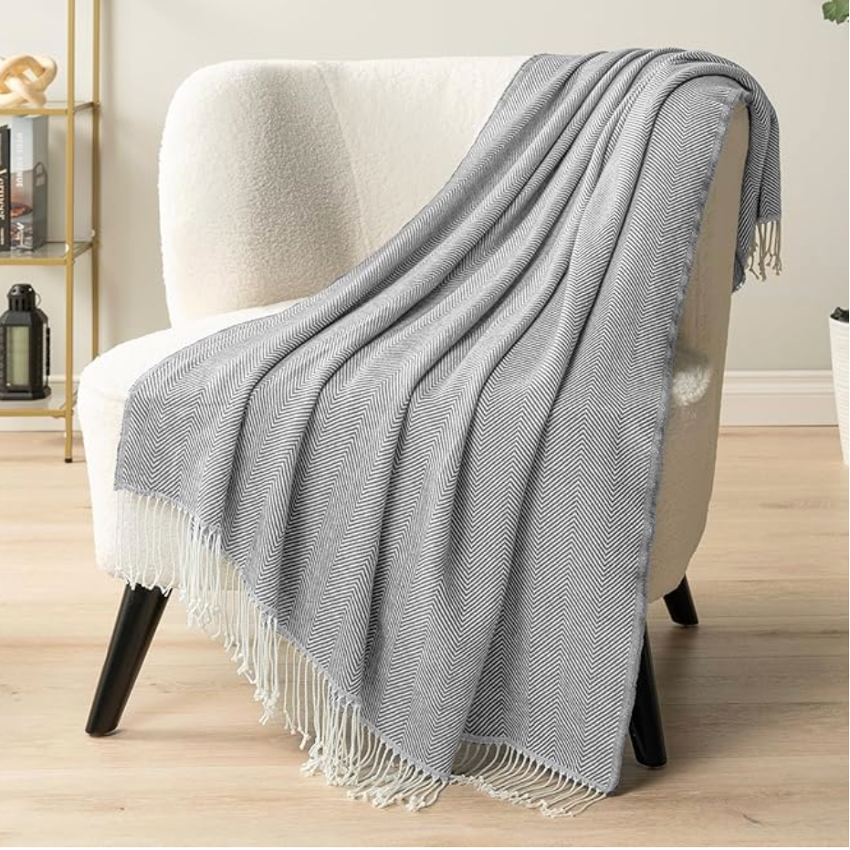 Gray throw blanket with white fringe draped over a white armchair in a room.