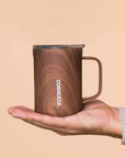 Hand holding a wooden-patterned mug with 'Corkcicle' branding against a beige background