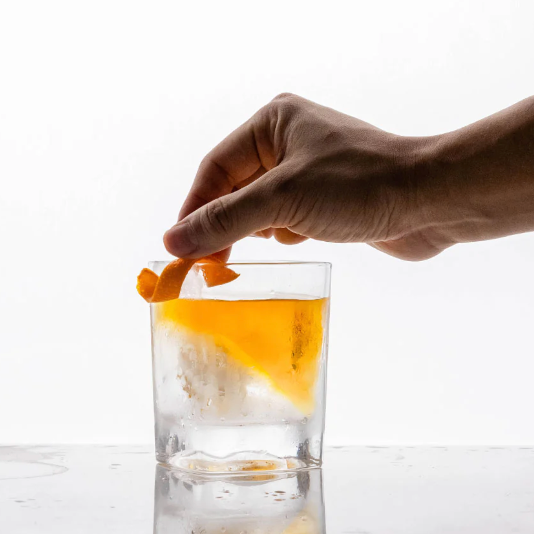 Hand placing an orange peel into a glass of alcohol on a white background