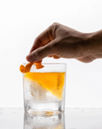 Hand placing an orange peel into a glass of alcohol on a white background