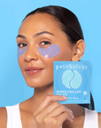 Woman wearing eye patches with a box of Patchology eye gels against a blue background