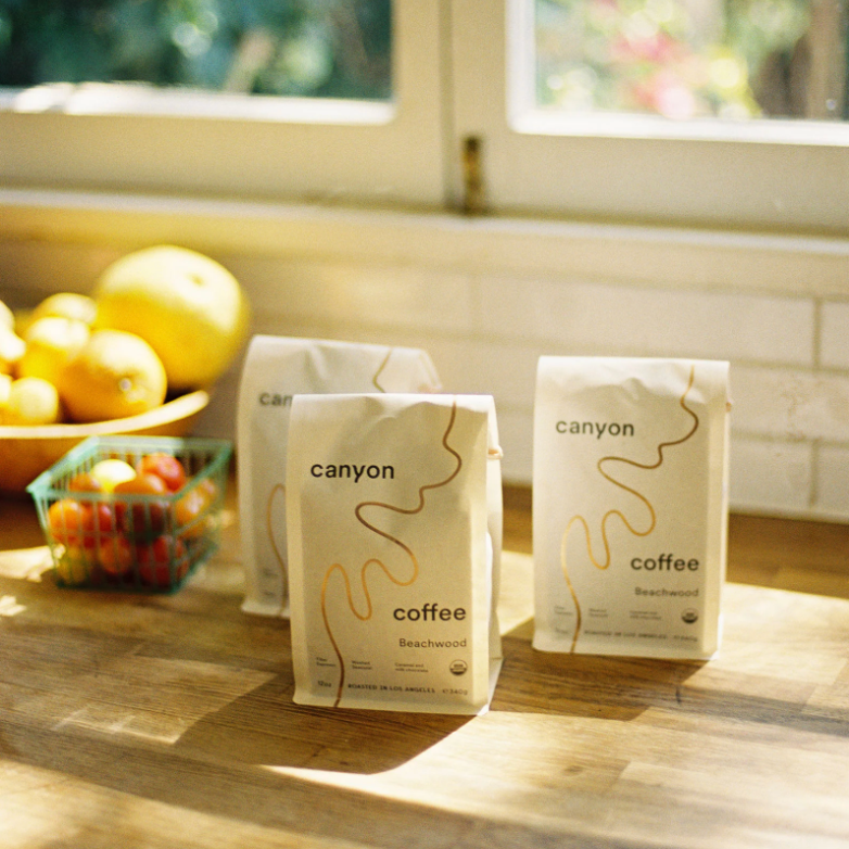 Canyon Coffee bags on a wooden surface with a window and fruit in the background