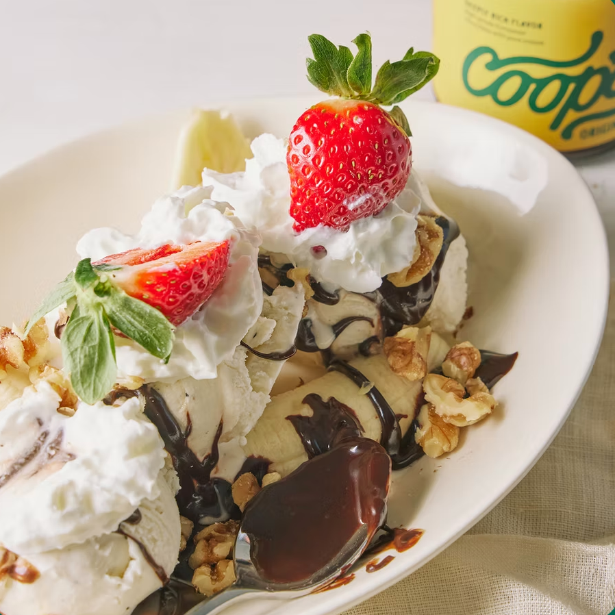 Ice cream sundae with whipped cream, strawberries, and chocolate sauce on a white plate, Coop logo in the background.