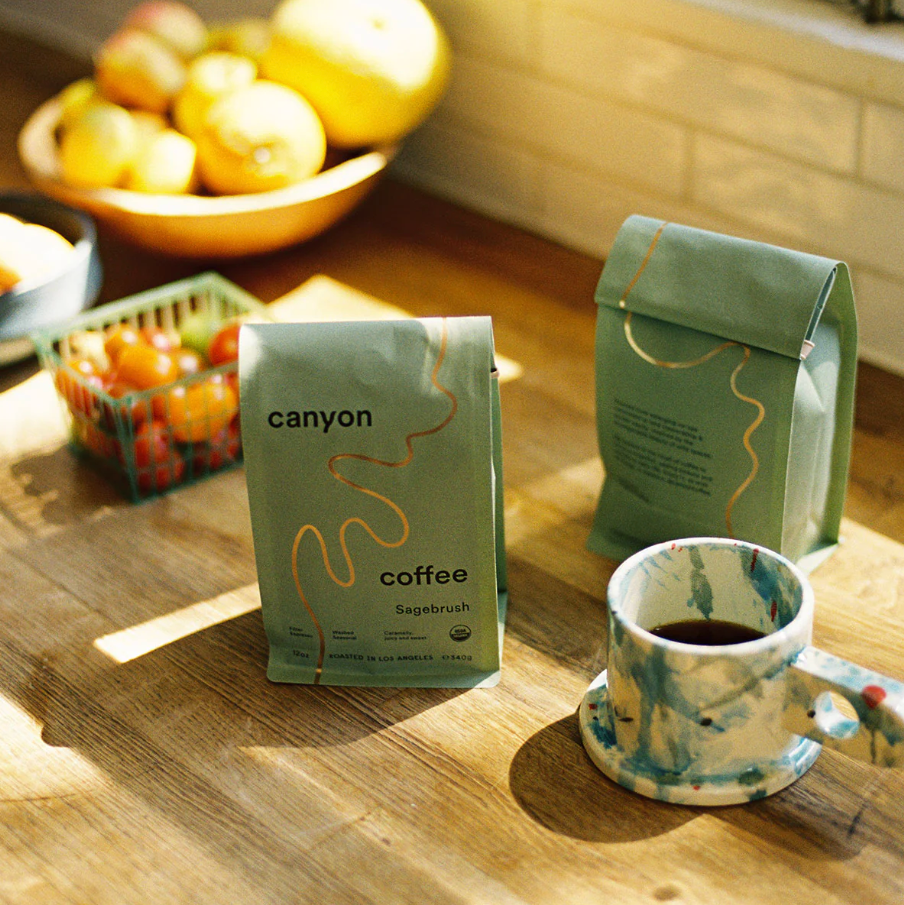 Two bags of 'canyon coffee' on a wooden surface with a cup of coffee and fruit bowl in the background.