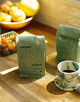 Two bags of 'canyon coffee' on a wooden surface with a cup of coffee and fruit bowl in the background.