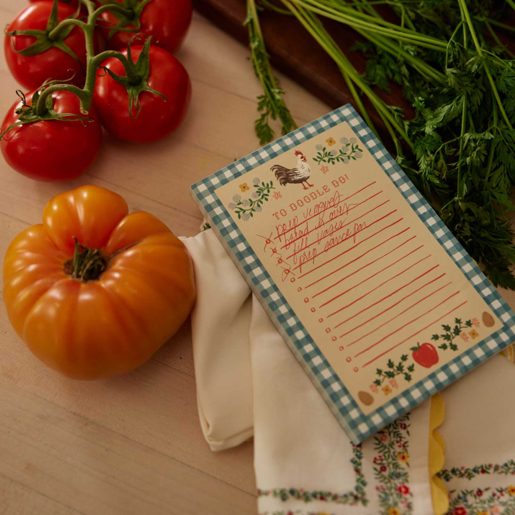 Notebook with a checkered border and rooster design on a wooden surface with tomatoes and herbs.