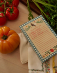 Notebook with a checkered border and rooster design on a wooden surface with tomatoes and herbs.