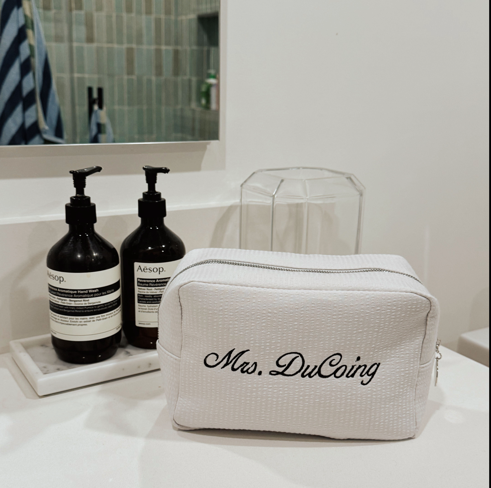 White toiletry bag with 'Mrs. DuCoing' branding on a bathroom counter with Aesop bottles.