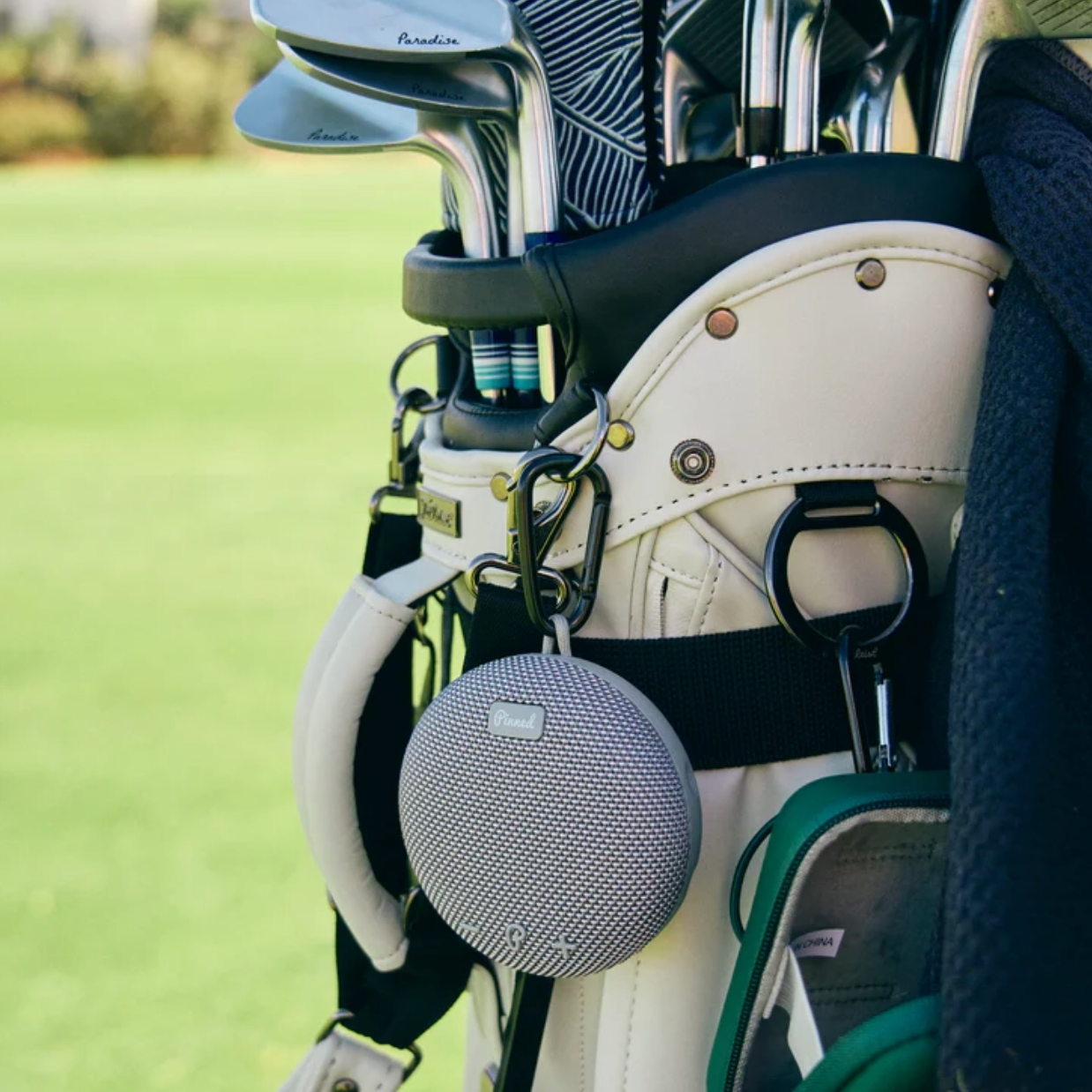 Close-up of a golf bag with golf clubs and a small speaker attached on a golf course.