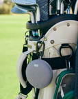Close-up of a golf bag with golf clubs and a small speaker attached on a golf course.