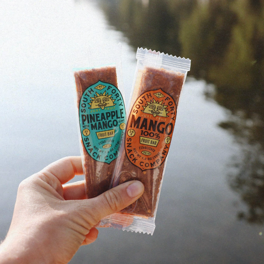 Two juice pouches held by a hand with a scenic background of water and trees.