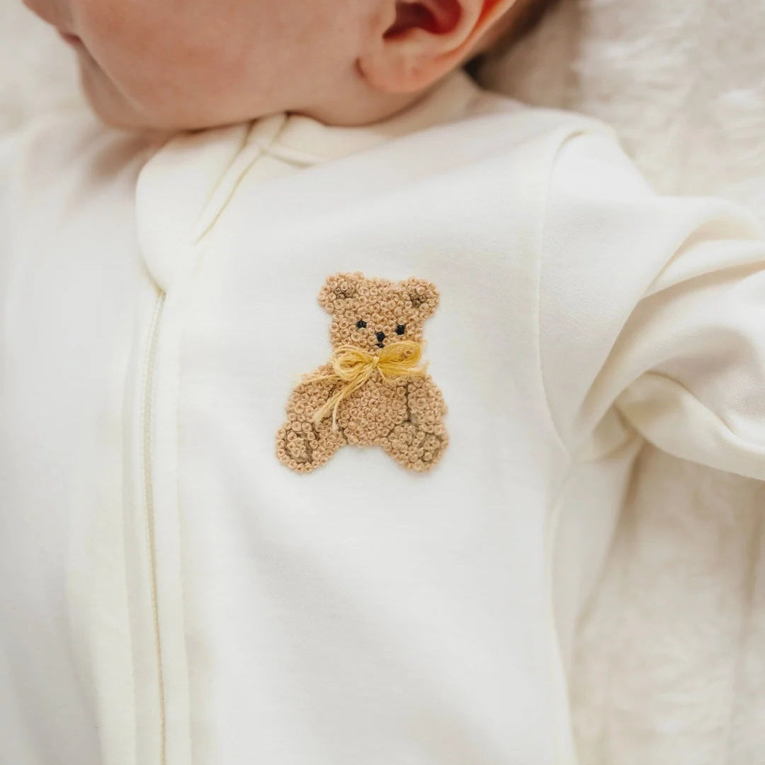 Close-up of a baby wearing a white zip-front bodysuit with a small embroidered teddy bear patch on the chest. The teddy bear is tan with a yellow bow, and the baby's ear and part of the face are visible.