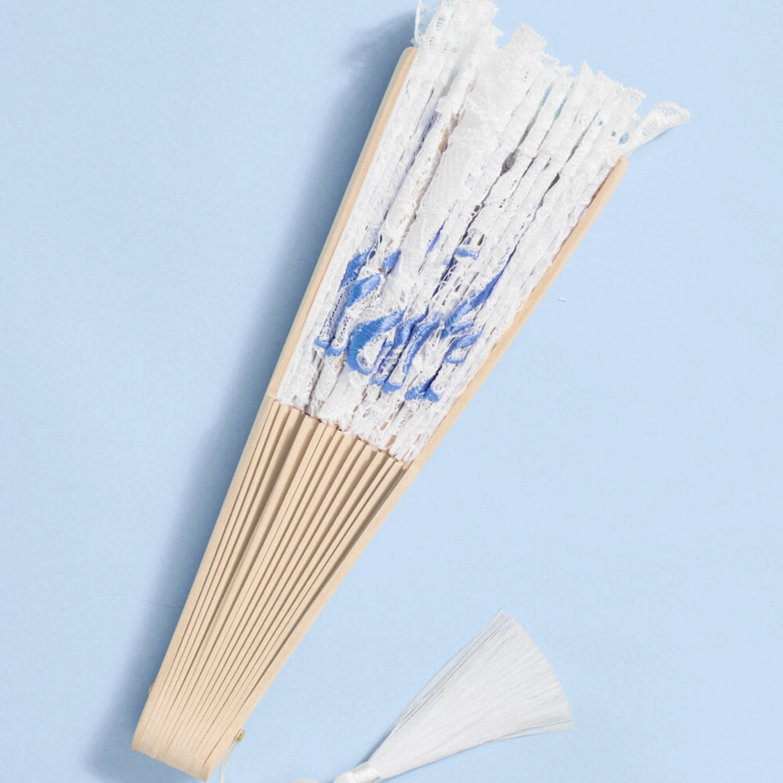A traditional folding hand fan with white paper ribs and blue floral printed panels, featuring a white tassel at the bottom, set against a light blue background.