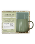 A tea gift set in a square box, featuring two packets of Tea With Tae matcha tea and a matte sage green ceramic mug with a matching lid, displayed on a white background.
