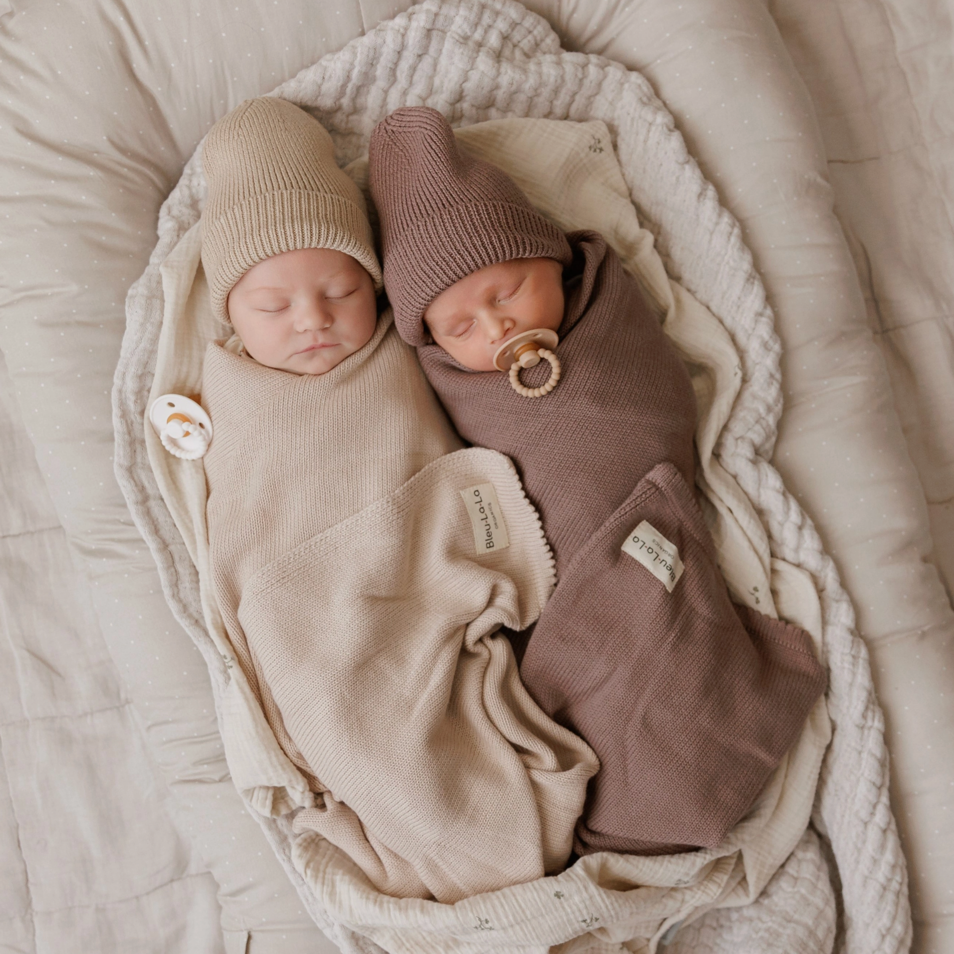 Two sleeping newborn babies swaddled in soft knit blankets and wearing knit hats, lying side by side in a bassinet. One swaddle is warm beige and the other is dusty mauve-brown, with matching pacifiers.
