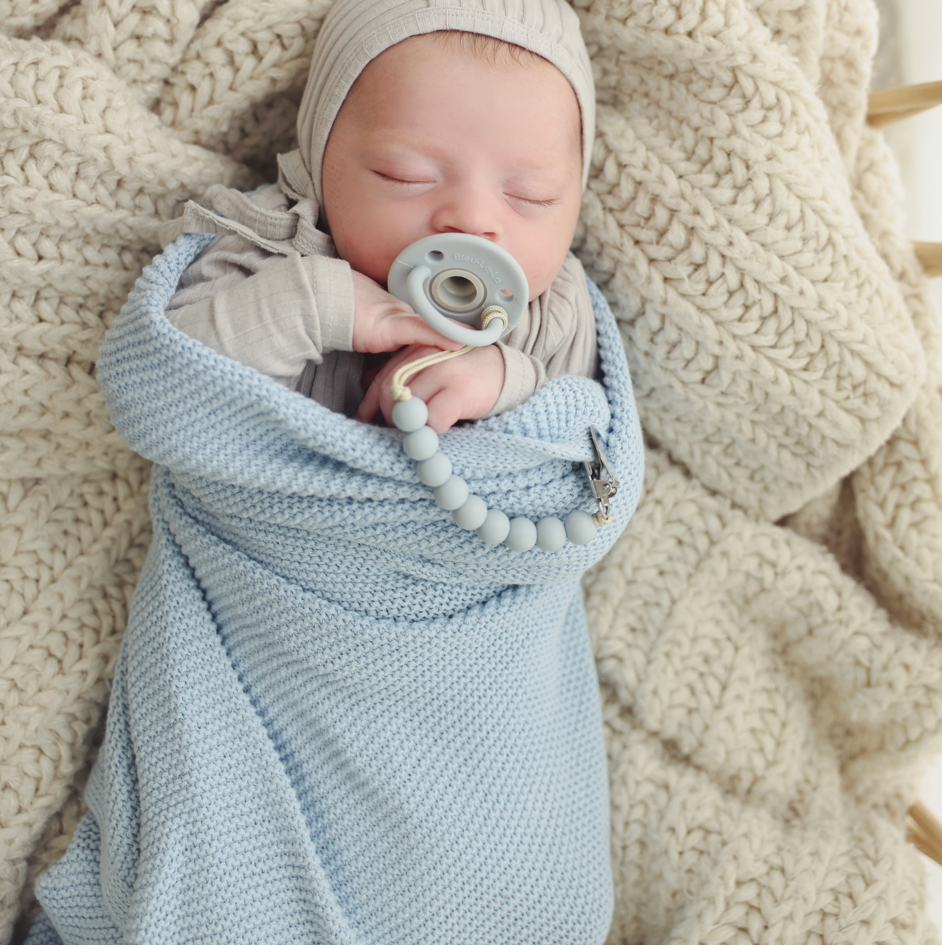 A sleeping newborn baby swaddled in a light blue knit blanket, wearing a beige outfit and bonnet, with a pacifier and beaded pacifier strap resting on their face, all set against a cozy cream-colored chunky knit blanket.