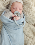 A sleeping newborn baby swaddled in a light blue knit blanket, wearing a beige outfit and bonnet, with a pacifier and beaded pacifier strap resting on their face, all set against a cozy cream-colored chunky knit blanket.