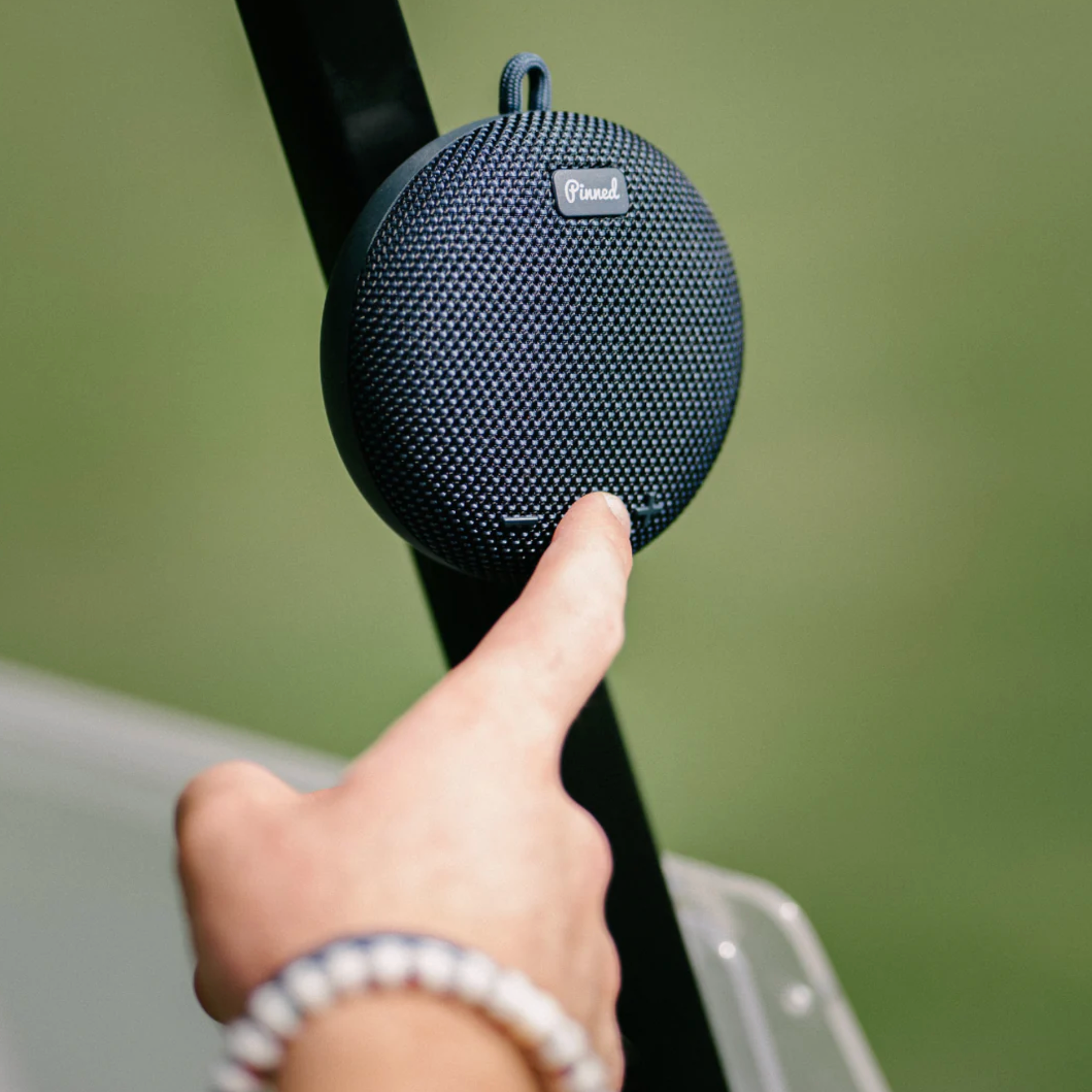 A close-up of a person's hand pressing a button on a round, dark blue Pund portable speaker mounted on a black pole against a soft-focus green background.