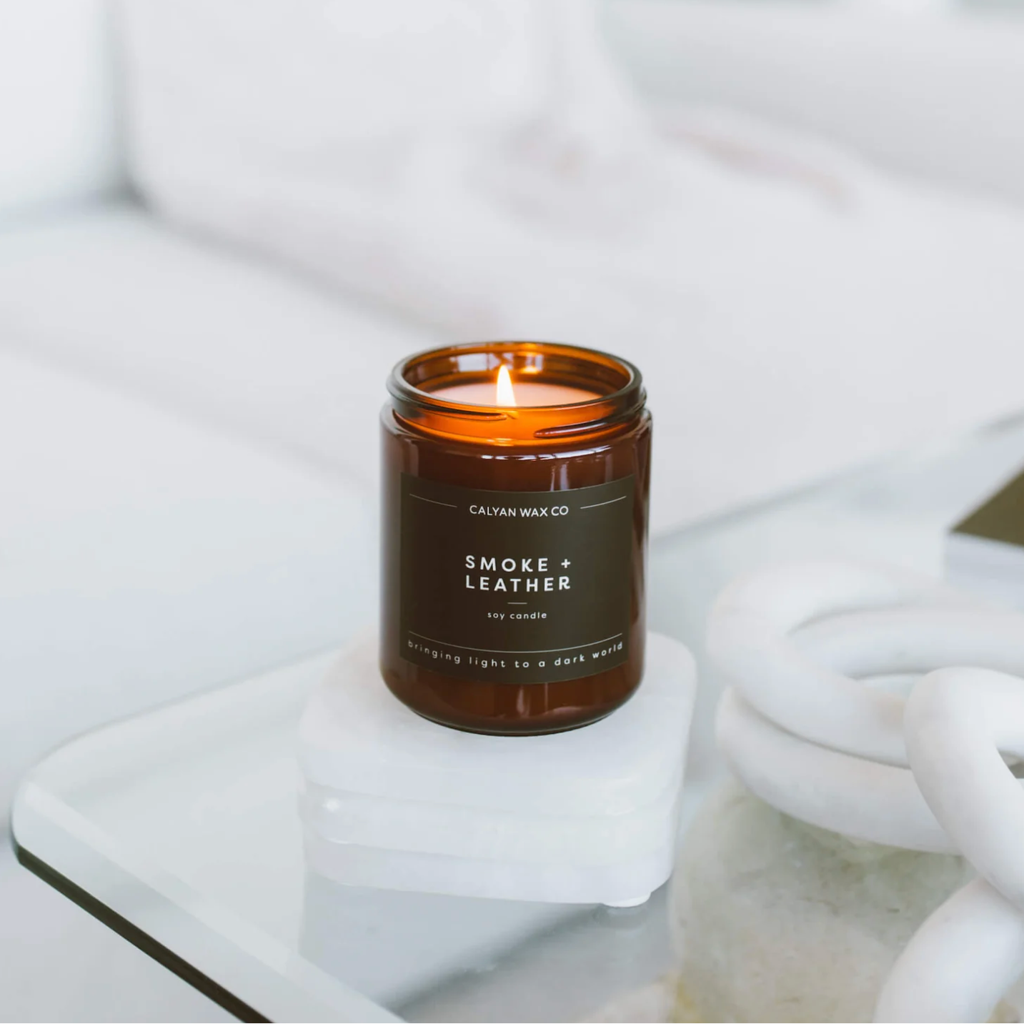 A lit amber glass jar candle labeled 'CALIFORNIA WAX CO. SMOKE + LEATHER' sits on a white marble coaster on a glass table in a bright, modern room.