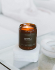 A lit amber glass jar candle labeled 'CALIFORNIA WAX CO. SMOKE + LEATHER' sits on a white marble coaster on a glass table in a bright, modern room.