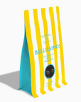 A standing pouch of Bellissimo dark chocolate caramels with yellow and white vertical stripes, a turquoise blue side panel, and a circular clear window showing dark brown chocolates inside, set against a plain white background.
