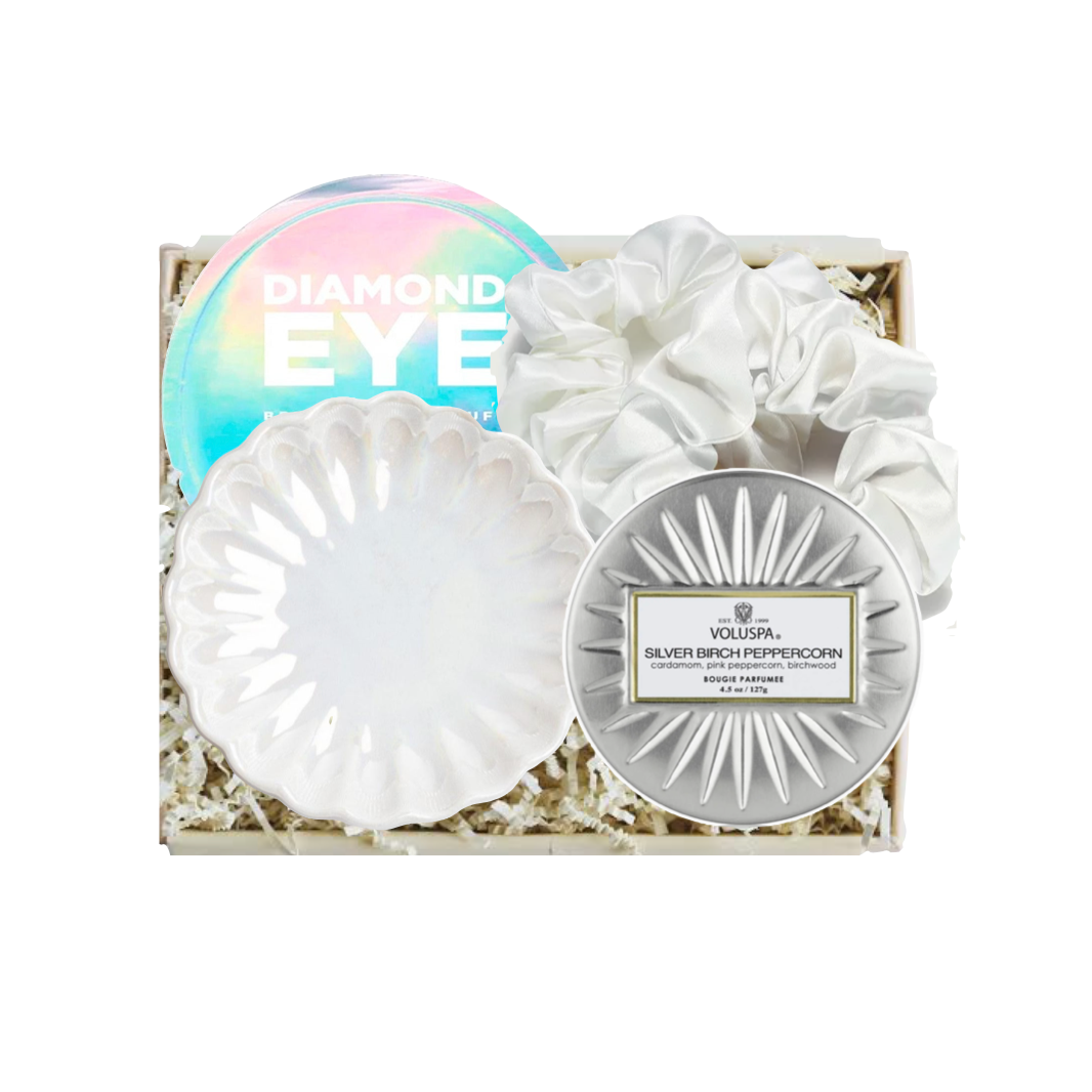 A boxed beauty gift set on a white background, containing a round iridescent eyeshadow palette labeled 'DIAMOND EYE,' a white scalloped makeup sponge, a white scalloped makeup brush, and a round silver compact with a label that appears to read Voluspa.
