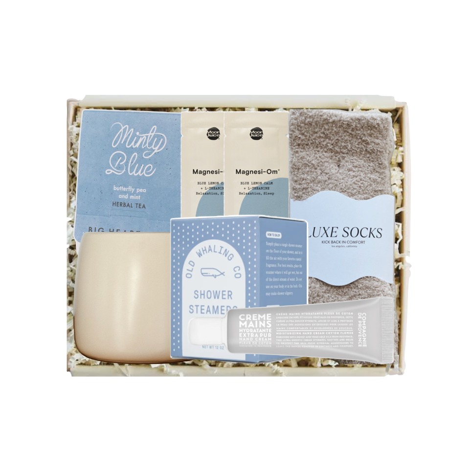 A curated self-care gift set in a light wood box with shredded paper filler, featuring a minty blue shower steam packet, cream-colored body wash, magnetic shower doors, plush gray socks, and a tube of cream hand cream.