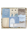 A curated self-care gift set in a light wood box with shredded paper filler, featuring a minty blue shower steam packet, cream-colored body wash, magnetic shower doors, plush gray socks, and a tube of cream hand cream.
