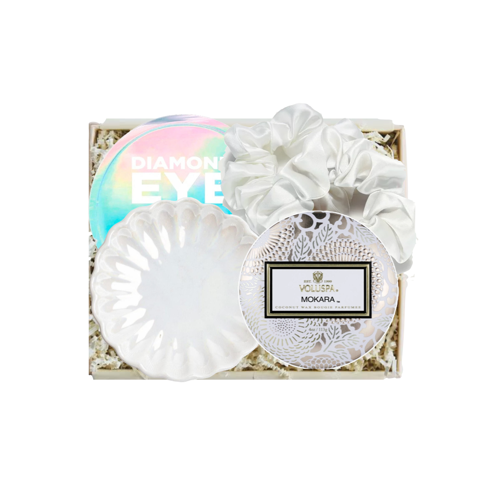 A white eye makeup gift set in a square box with shredded paper filler, featuring a white scalloped eyeshadow sponge, a white fabric scrunchie, a round white doily-style compact with a Nora.