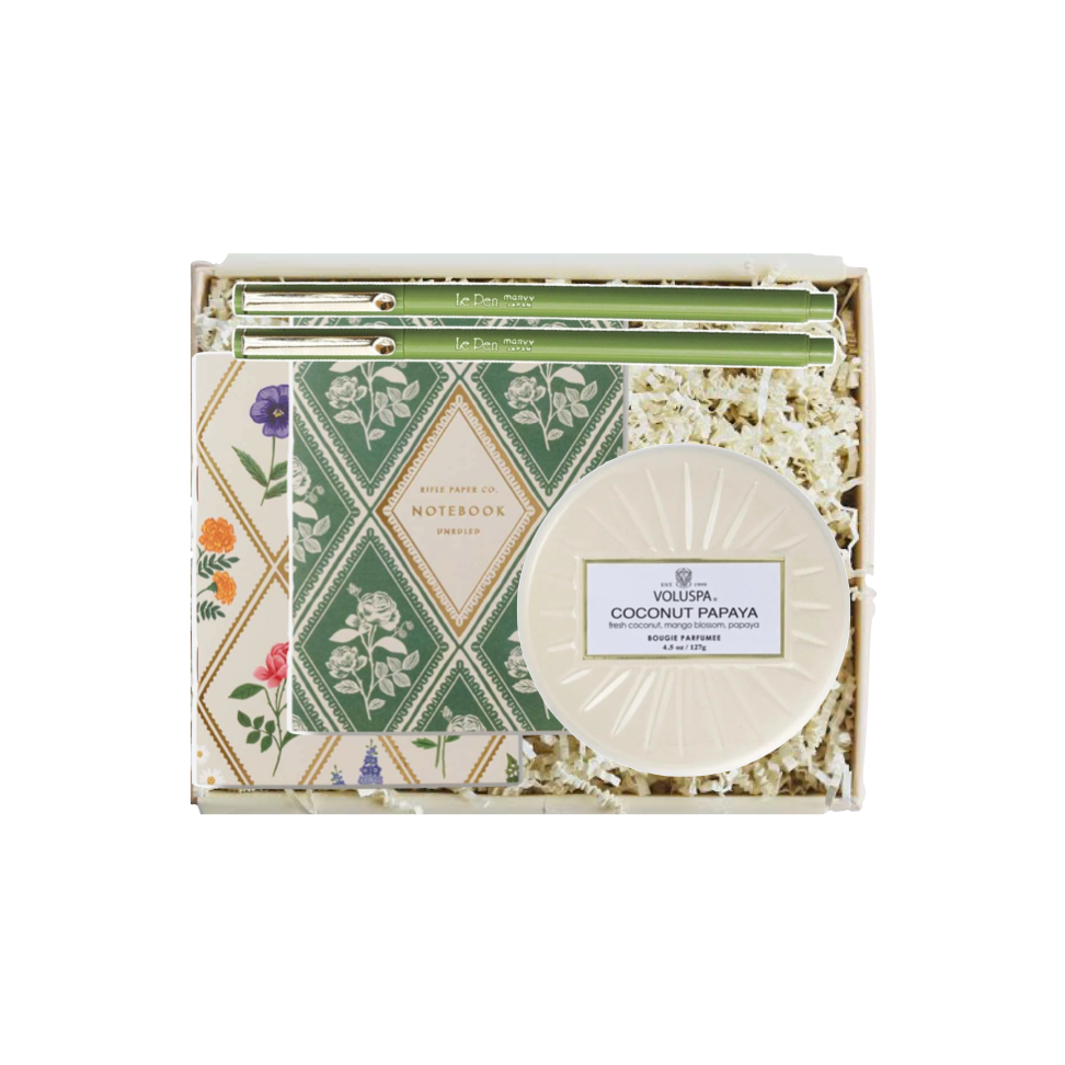 A boxed gift set from Volupta featuring a round coconut papaya soap, a patterned notebook, and green pens arranged in white crinkle paper filler inside a clear gift box.