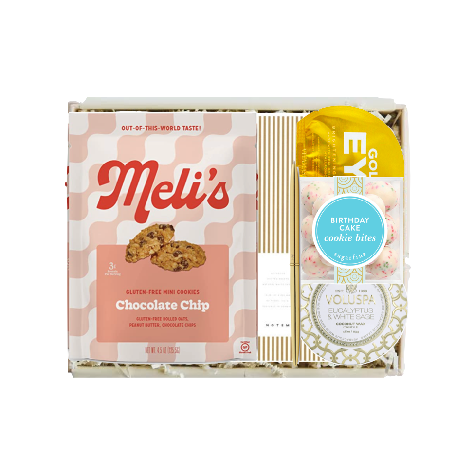 A curated gift box set from Meli's and Voluspa, featuring a box of Chocolate Chip cookie bites, a Voluspa Cocoon of Voluspa Garcinia & White Sugar cookies, and a yellow and blue birthday cake cookie bite packet, arranged in a white box with shredded paper.