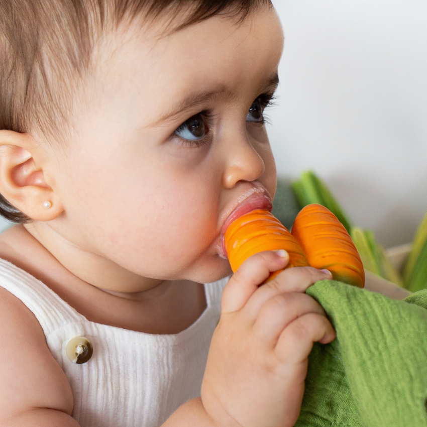 A baby wearing a white sleeveless top is shown in a close-up, holding and biting an orange carrot-shaped teething toy attached to a green fabric strap.