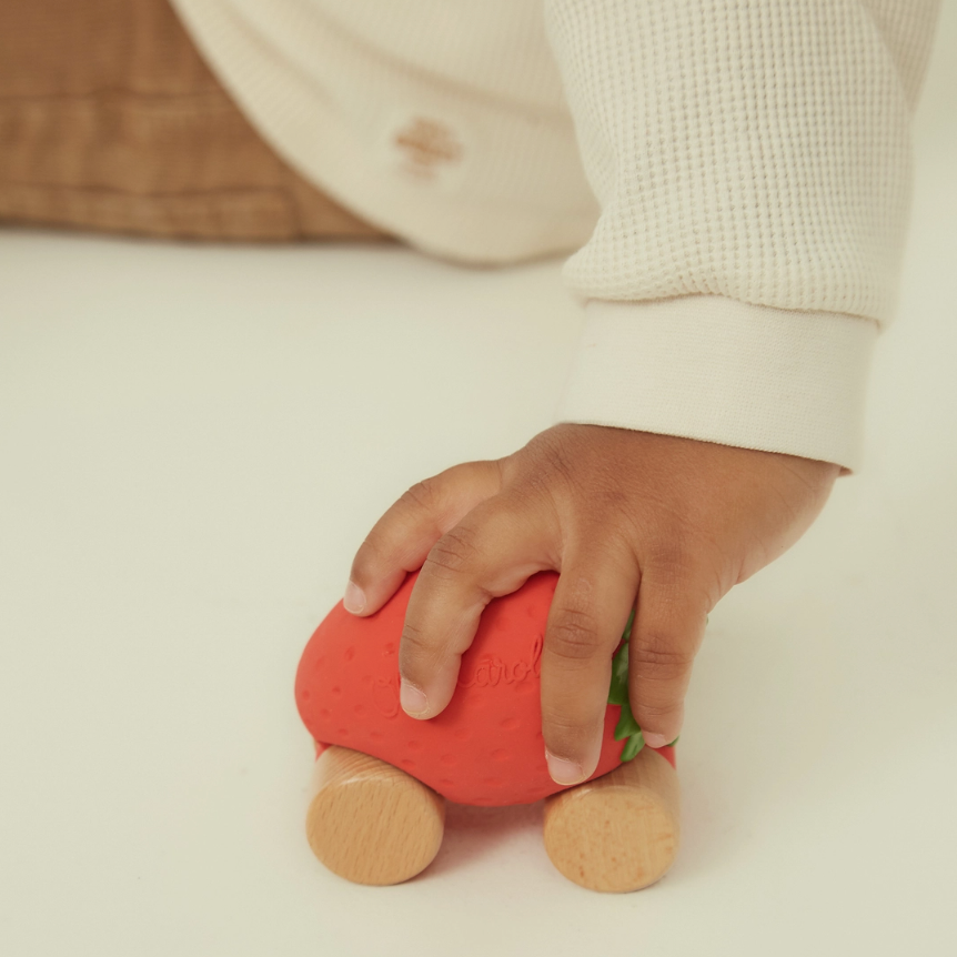 A child's hand pushes a small wooden toy car shaped like a strawberry on a clean white floor. The toy has a red body, green leaf detail, and natural wood wheels.