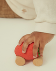 A child's hand pushes a small wooden toy car shaped like a strawberry on a clean white floor. The toy has a red body, green leaf detail, and natural wood wheels.