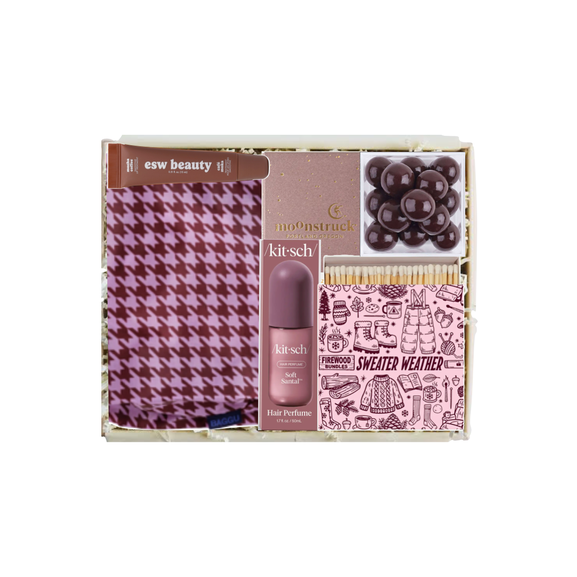 A boxed beauty gift set on a white background, containing a pink and purple houndstooth scarf, a bottle of Kitsch hair perfume, a pink box with a gold logo, a tray of dark brown chocolate-covered candies, and a pink patterned packet labeled 'SWEATER WEATHER'.