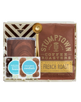 A coffee gift set in a clear window box package, featuring a brown Stumptown French Roast coffee bag, a glass carafe with dark roasted coffee, a gold spoon, and two small cold brew packets, arranged on white paper shred.