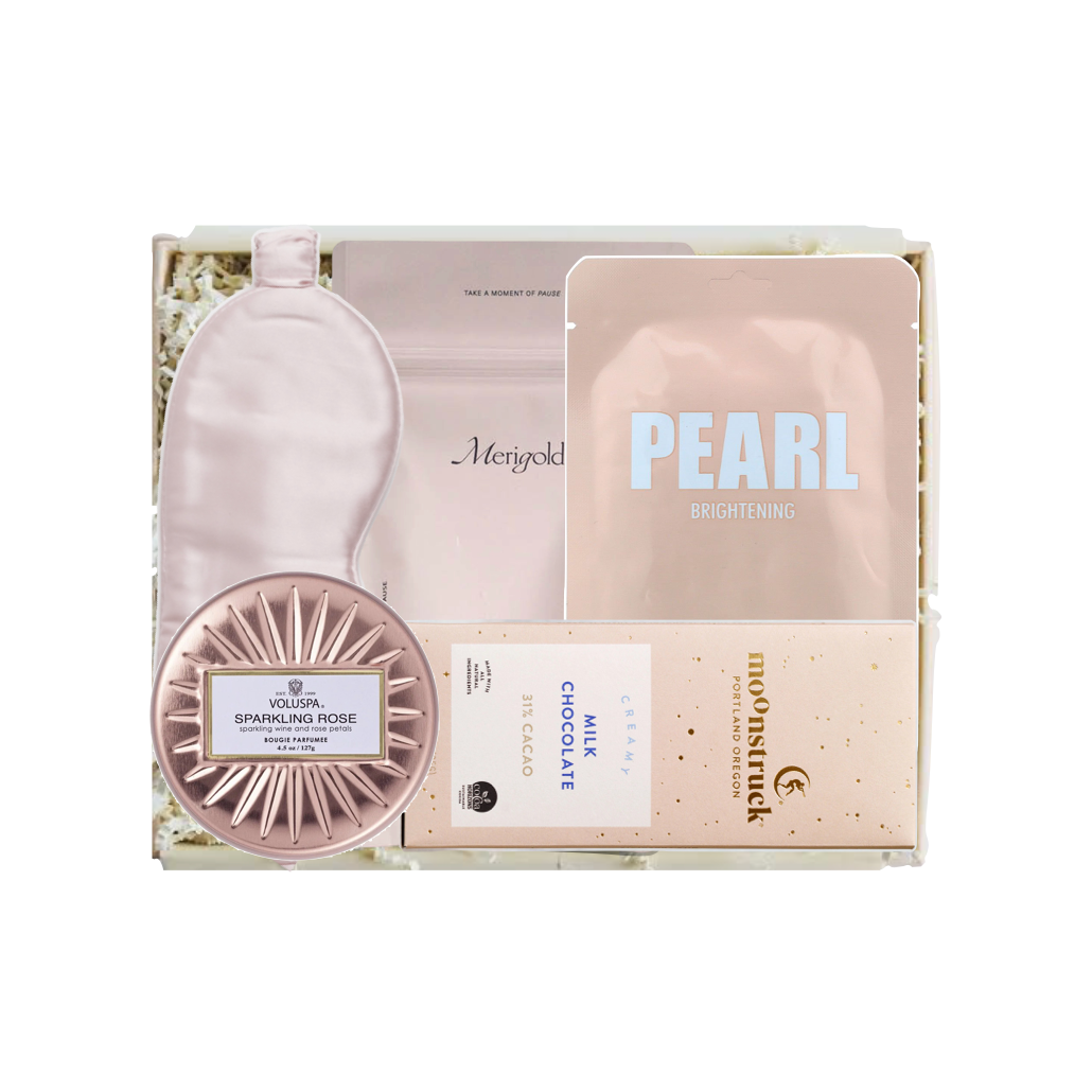 A curated beauty gift set in a white box, featuring a pale pink sleep mask, a pink jar labeled 'SPARKLING FACE GEL', a pink pouch labeled 'PEARL BRIGHTENING', a chocolate bar labeled 'CHOCOLATE', and a small round compact.