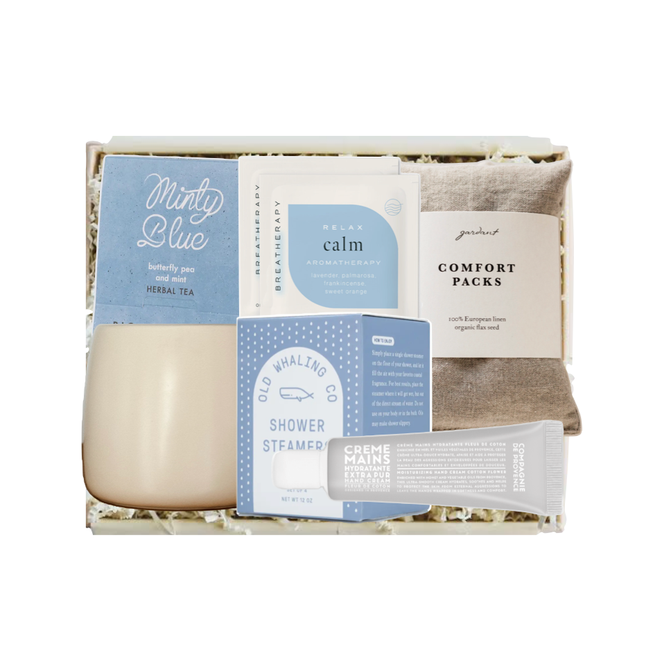 A curated wellness gift set in a white box with shredded paper filler, featuring a beige candle, blue and white packaging cards, a comfort pack pouch, and a gray cream packet on a white background.