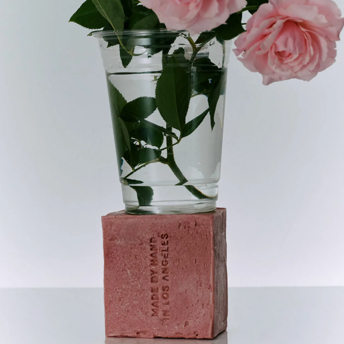 A clear plastic cup filled with water and rose stems, placed on top of a pink rectangular block that reads 'MADE BY MARY IN LOS ANGELES,' with pink roses extending from the cup against a clean white background.