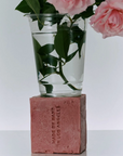 A clear plastic cup filled with water and rose stems, placed on top of a pink rectangular block that reads 'MADE BY MARY IN LOS ANGELES,' with pink roses extending from the cup against a clean white background.