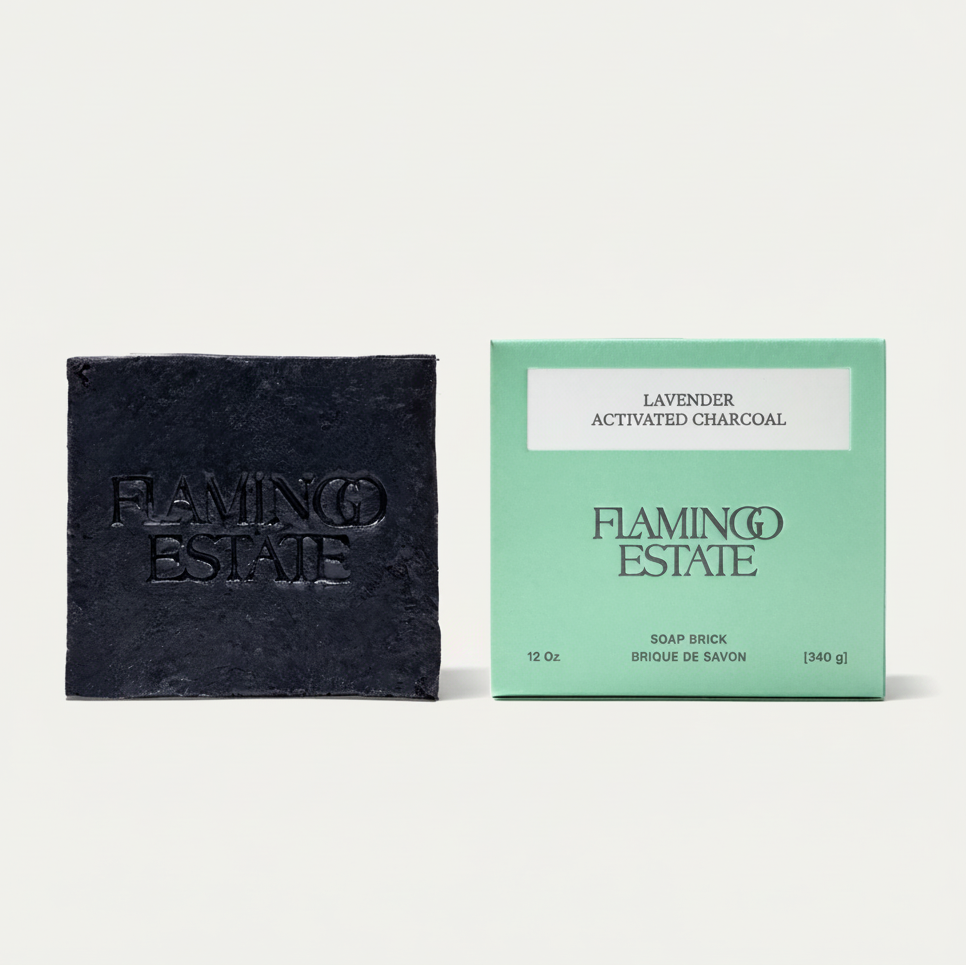 A dark charcoal soap brick next to its mint green packaging box, both embossed with the brand name 'FLAMINGO ESTATE'. A bright green circular badge in the top right corner reads 'NEW LOOK SAME FORMULA'.